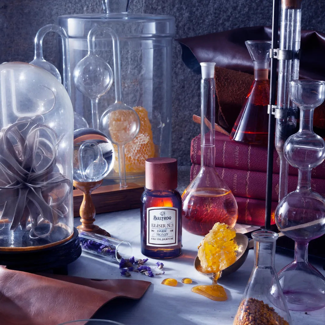 Bullfrog Elisir N.3 Dark Honey EdP parfymflaska omgiven av laboratorieutrustning, böcker och honung - en doft med läder, ros och bärnsten.