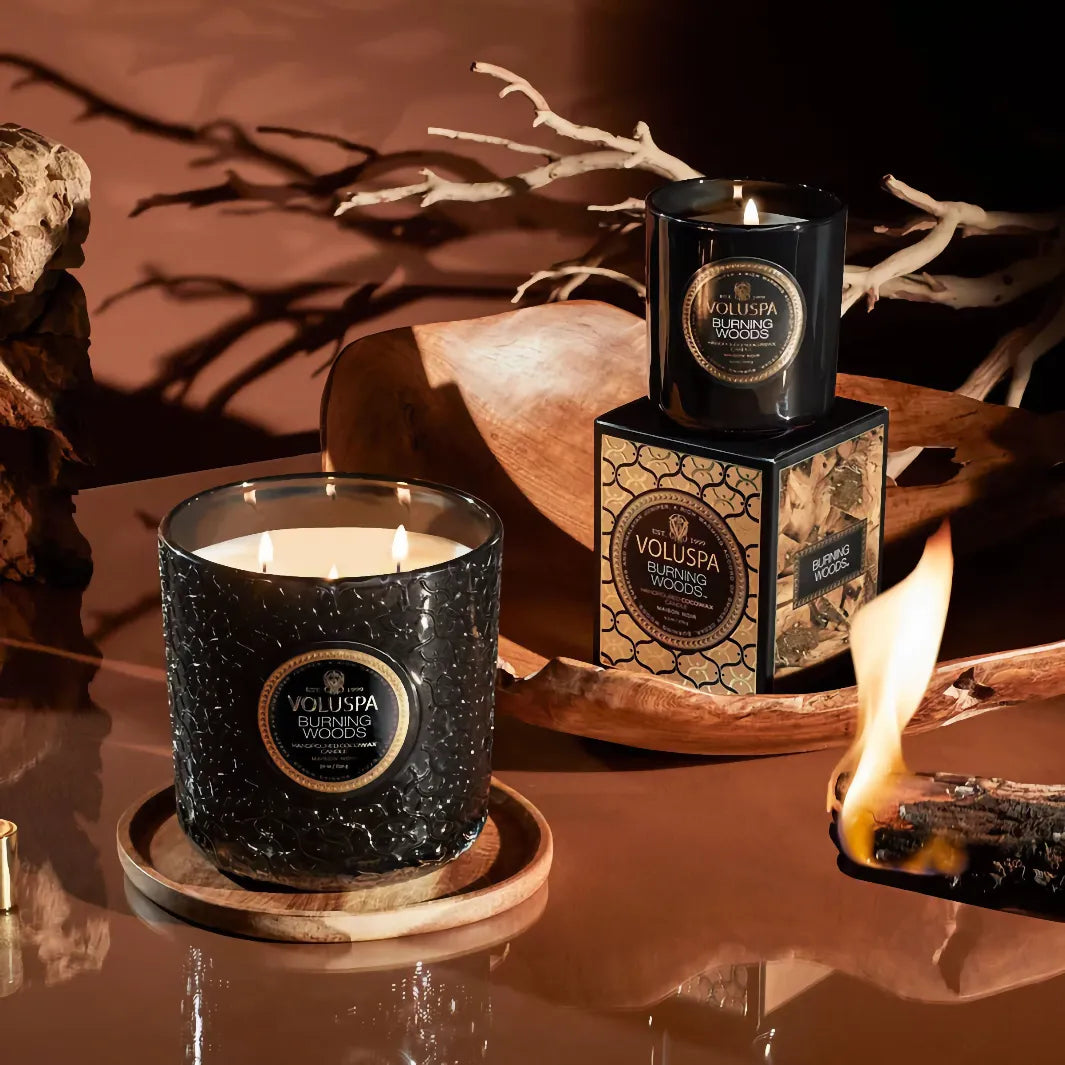 Voluspa Burning Woods doftljus i stämningsfull miljö med brinnande ljus, dekorativa trädetaljer och presentförpackning